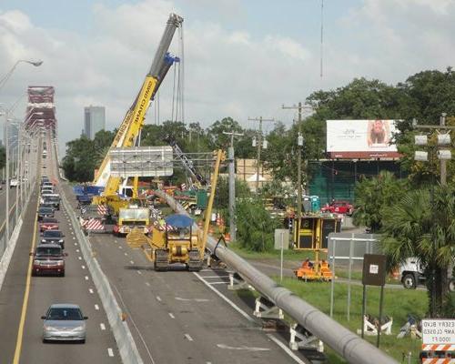 Very long pipe on stands along a highway full of cars. Large tractors, a crane, and other equipment there for the installation.